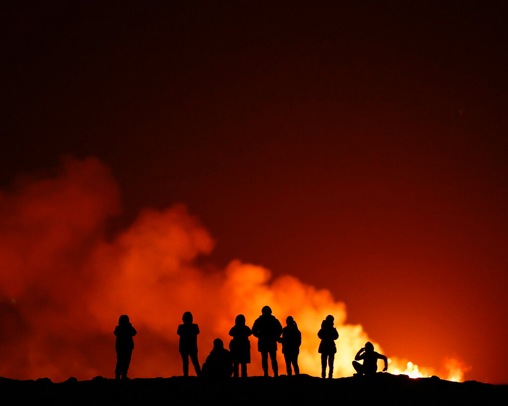 A group of people are silhouetted against the glow of the fire and smoke of an active volcano at night.