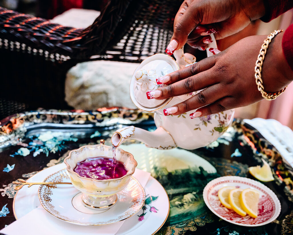 A woman’s hands with long nails pours a cup of purple tea from a delicate teapot.