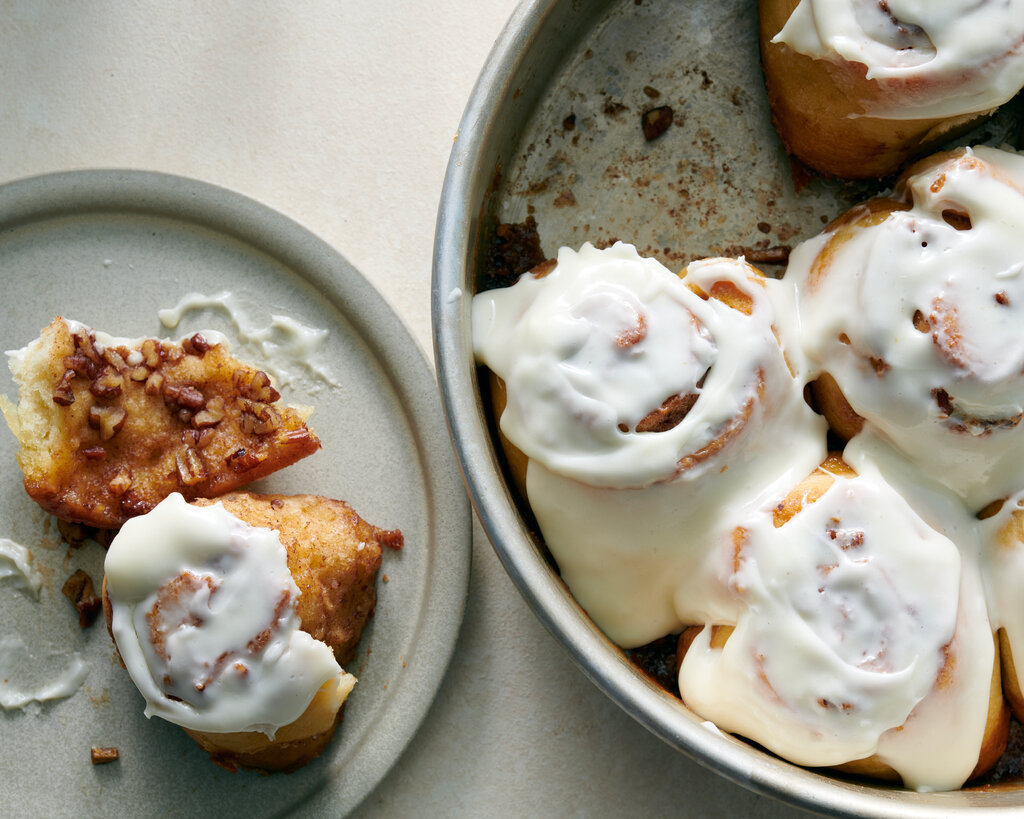 A plate with two round, icing-covered cinnamon rolls sits next to a pan with four more rolls.