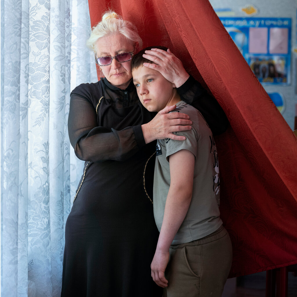 A grandmother embraces her grandchild near a window with a red curtain behind them. 