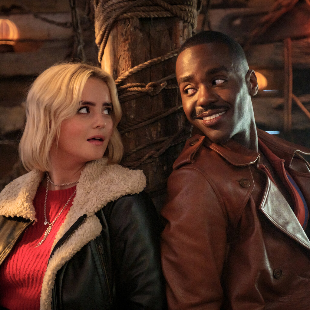 A woman in a leather jacket and read sweater has her back to a wooden pole and looks to her left at a smiling man in a maroon jacket also with his back to the pole.