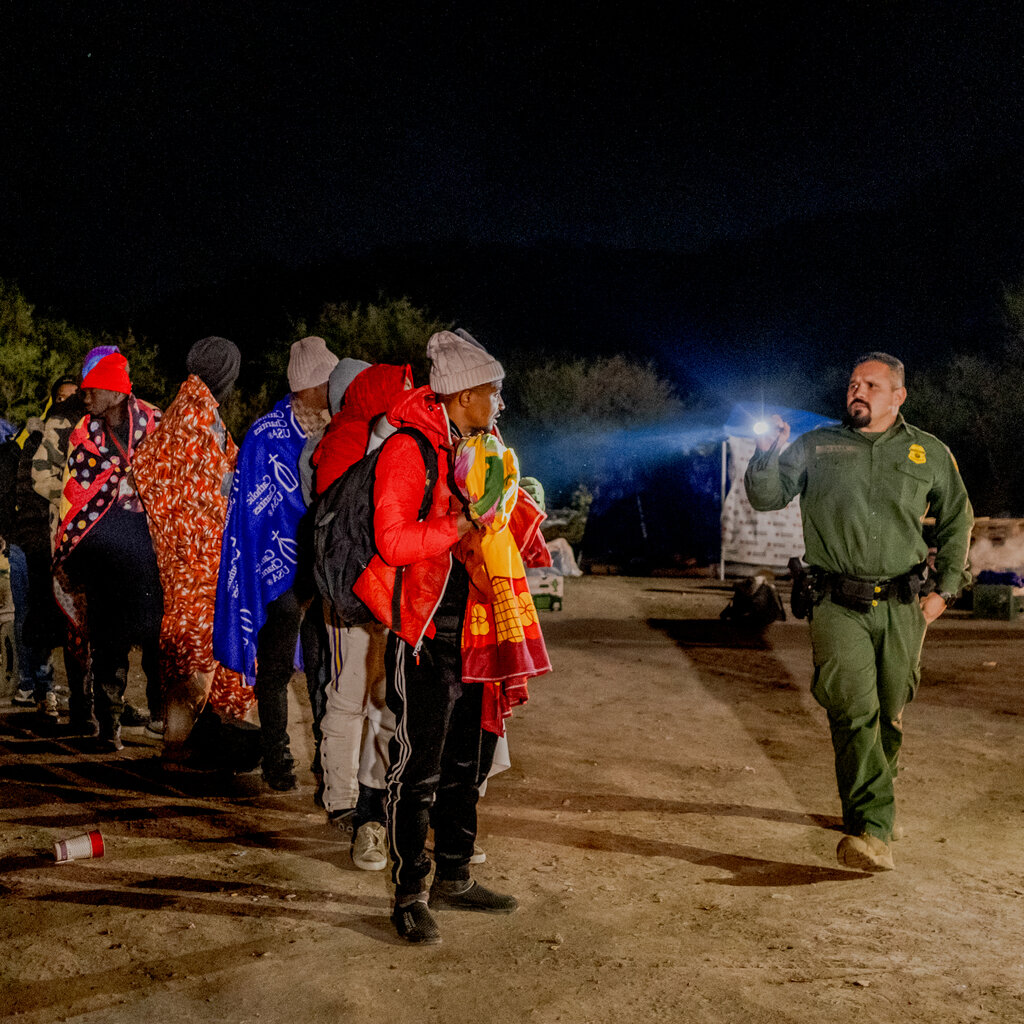 A Border Patrol agent in a green uniform points his flashlight at a group of people standing outside in blankets and warm clothes.
