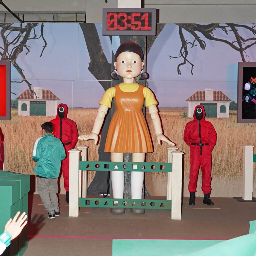At a “Squid Game” live event, people run around in front of a replica of the show’s large killer doll; people dressed like the show’s henchmen stand on either side.