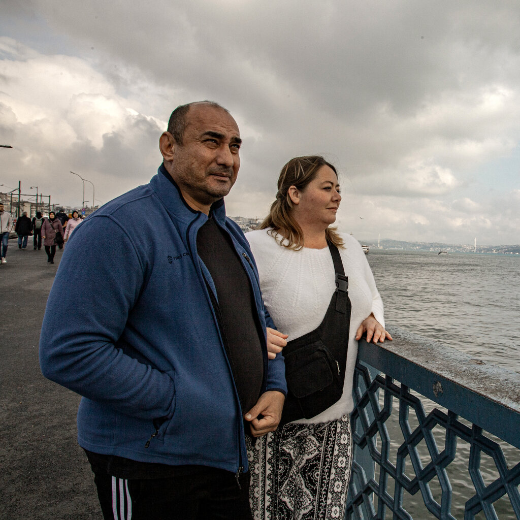 A middle-aged man wearing a blue fleece and a woman wearing a white sweater stand pensively on a walkway by the water. 