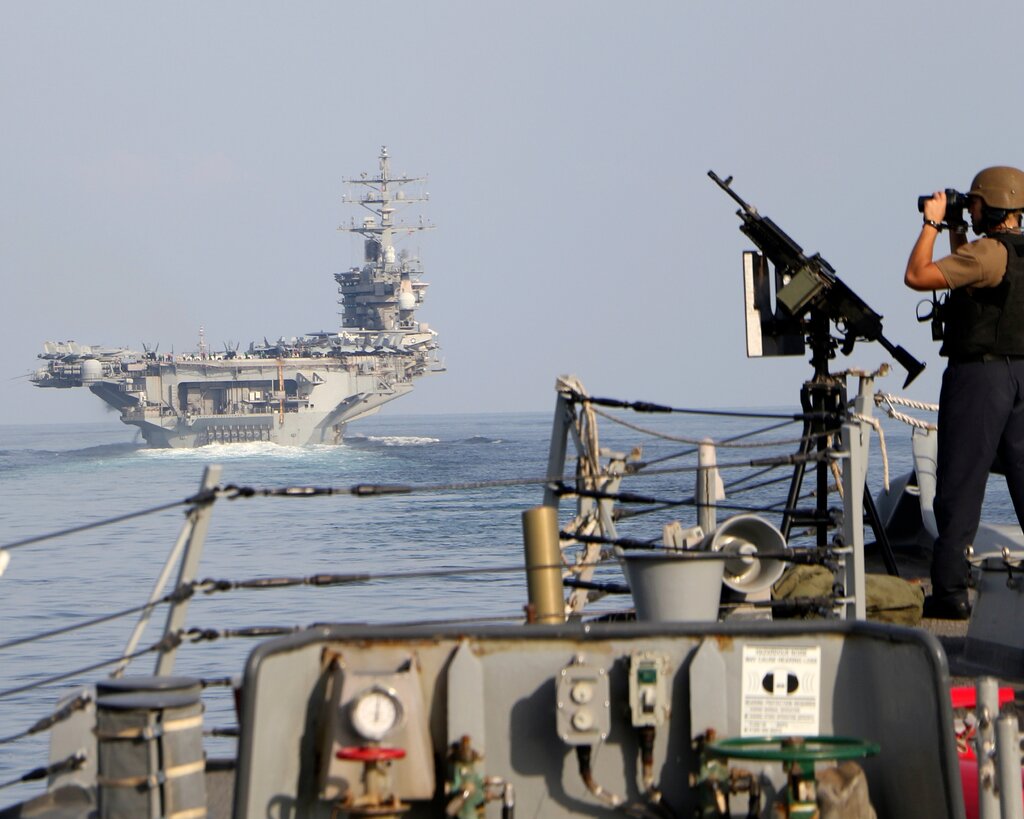 A man stands on the deck of a ship holding binoculars. An aircraft carrier is seen in the distance.
