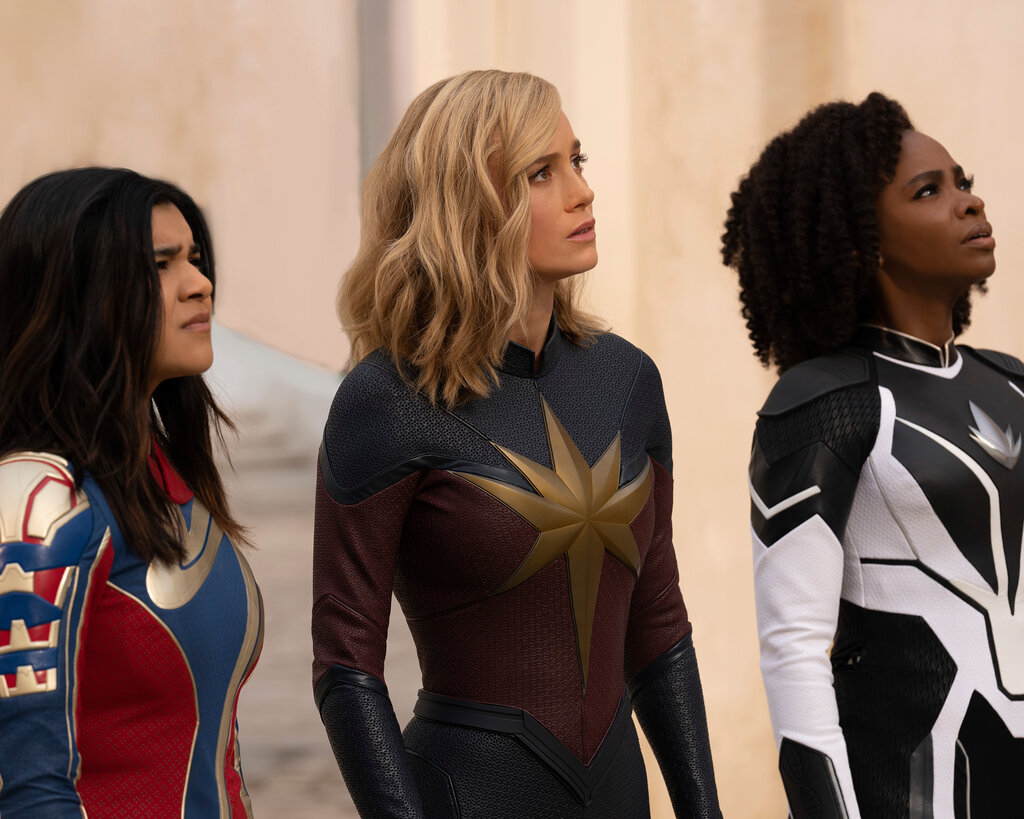 Three women — Iman Vellani, Brie Larson and Teyonah Parris — in Marvel character superhero suits look up toward the sky.