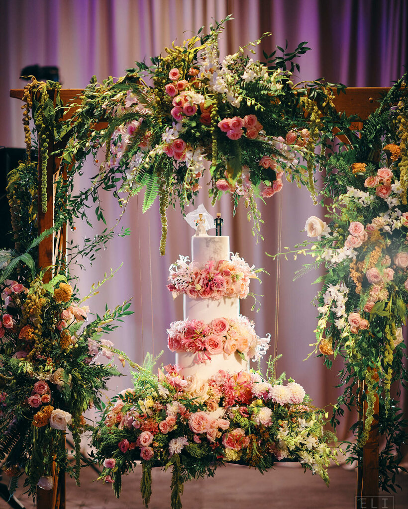An elaborate three-tiered cake with white frosting covered in pink flowers and topped with bride and groom figurines.