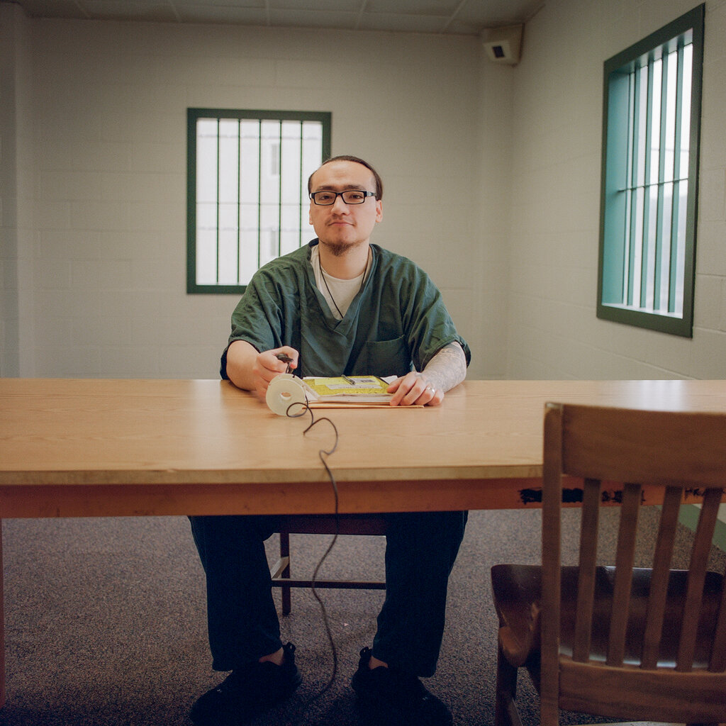 A man wearing a green prison uniform, sitting at a wooden desk in a room with barred windows.