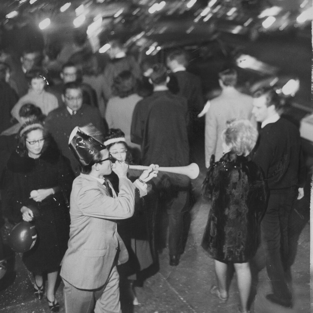 A black-and-white photo of a crowded street, with a person in a party hat blowing into a plastic horn.