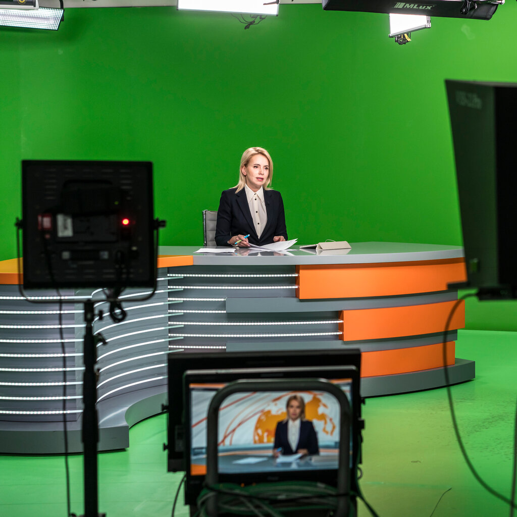 A woman sits on a desk in front of a green screen. Cameras are recording her. 