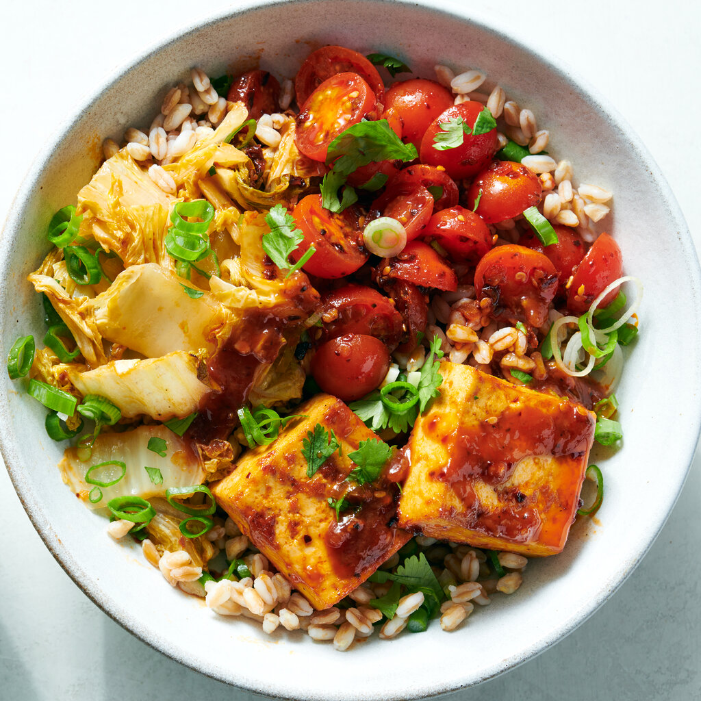 Two white bowls hold cooked grains topped with marinated cherry tomatoes and roasted sweet chile tofu and cabbage.