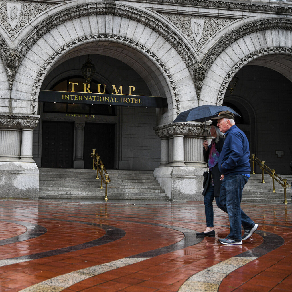 Two people holding an umbrella walk in front of a building, which has a sign that reads "Trump International Hotel."