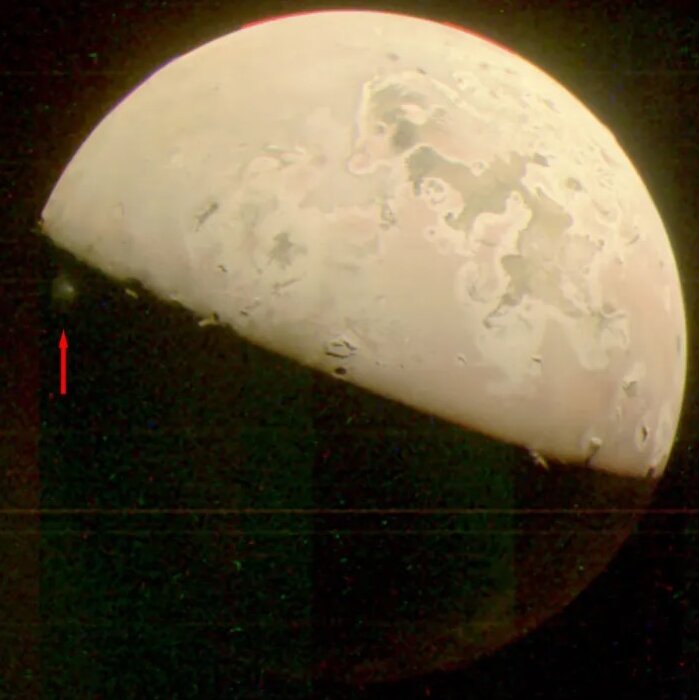 A view of Io, which appears tilted and half bright, half dark, and has a little whitish plume on an edge of its dark side, with a red arrow superimposed to point it out.