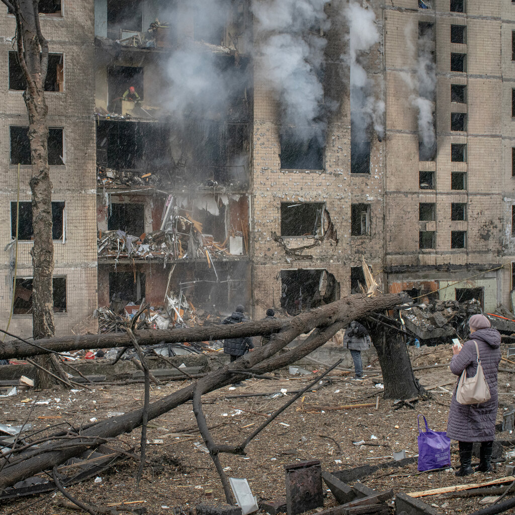 Smoke rises from a residential building in Kyiv after an airstrike. A person stands in the foreground looking at their phone.