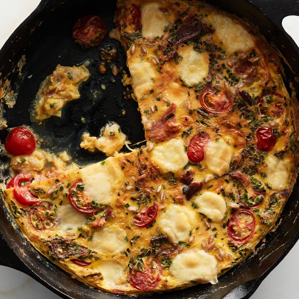 A cast-iron skillet holds a cheesy frittata with cherry tomatoes and dollops of cheese.
