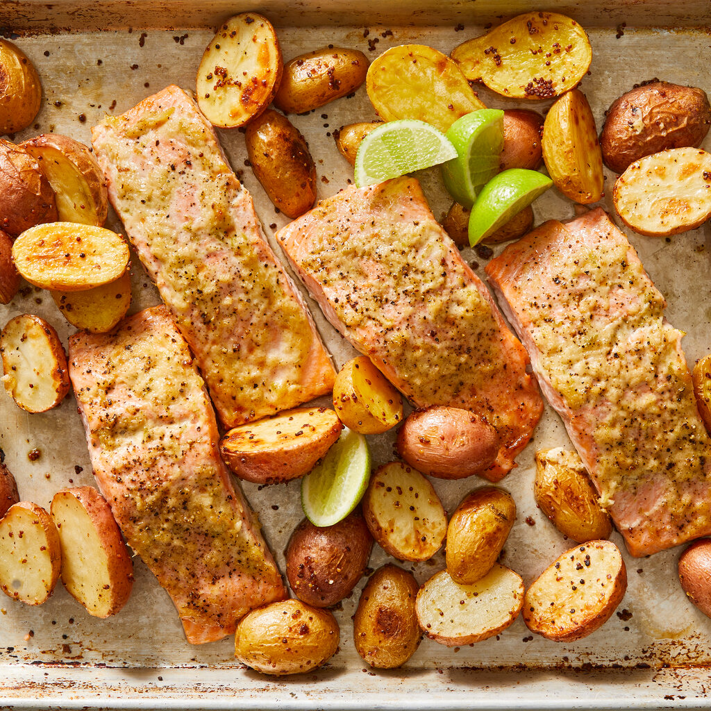 A sheet pan holds four salmon fillets roasted with horseradish and lime zest, surrounded by sliced and browned mustard potatoes and a handful of lime wedges.