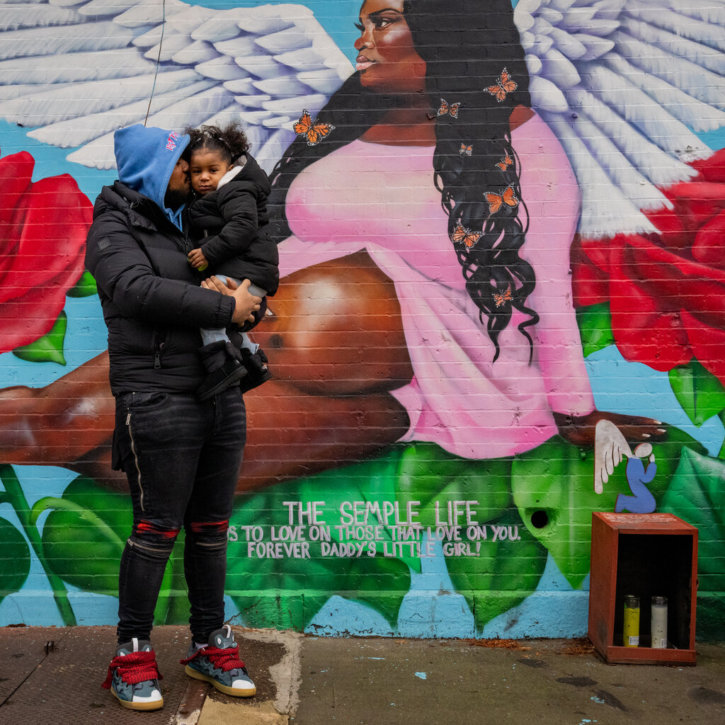A father dressed in a black coat holds a little girl, also in a black coat, in front of a mural depicting her mother, who is wearing a pink dress that shows her bare pregnant belly.