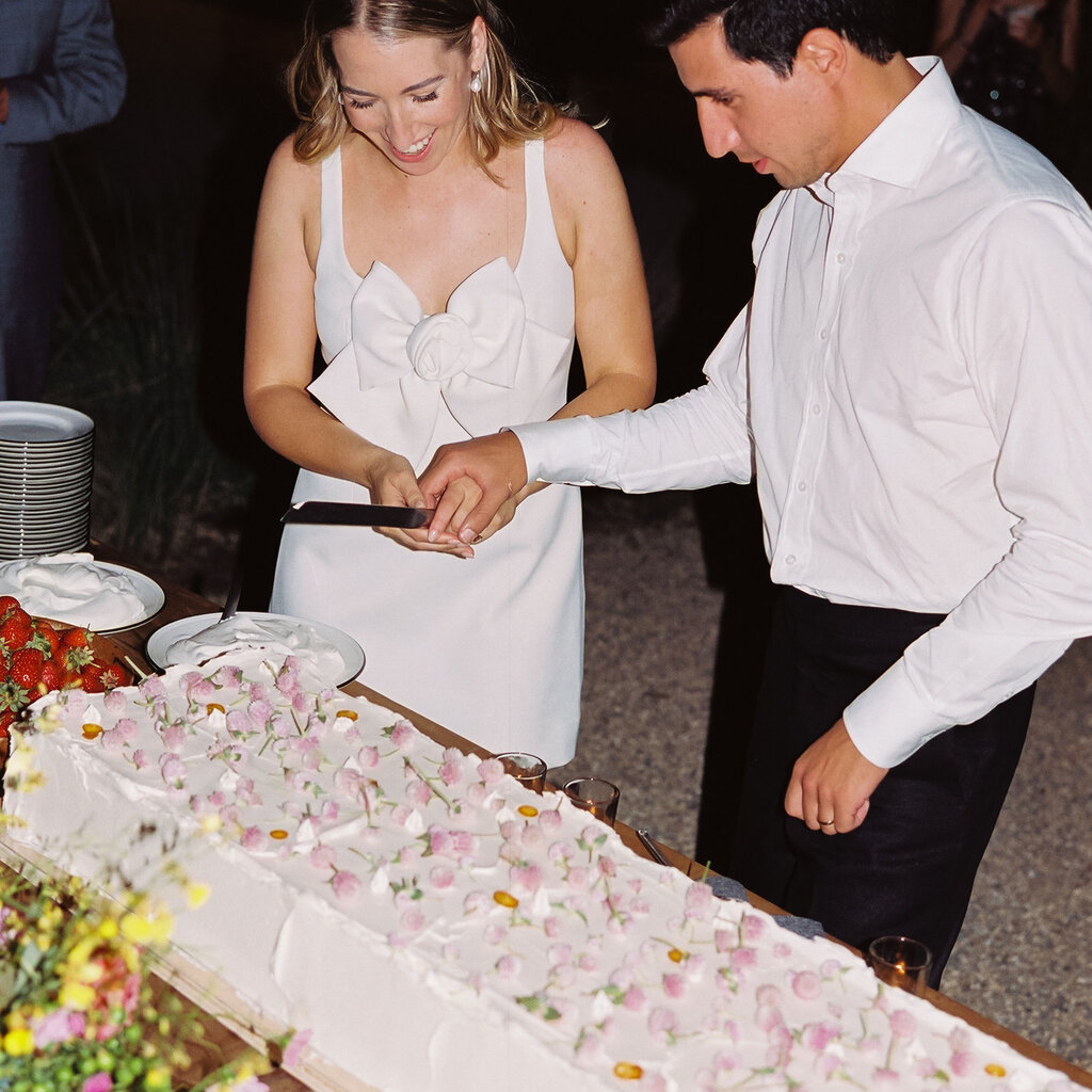 Rachel Karten, wearing a white dress with a large bow and rosette on the front, smiles as she and her spouse prepare to cut into a long sheet cake covered in white frosting and pink flower petals.