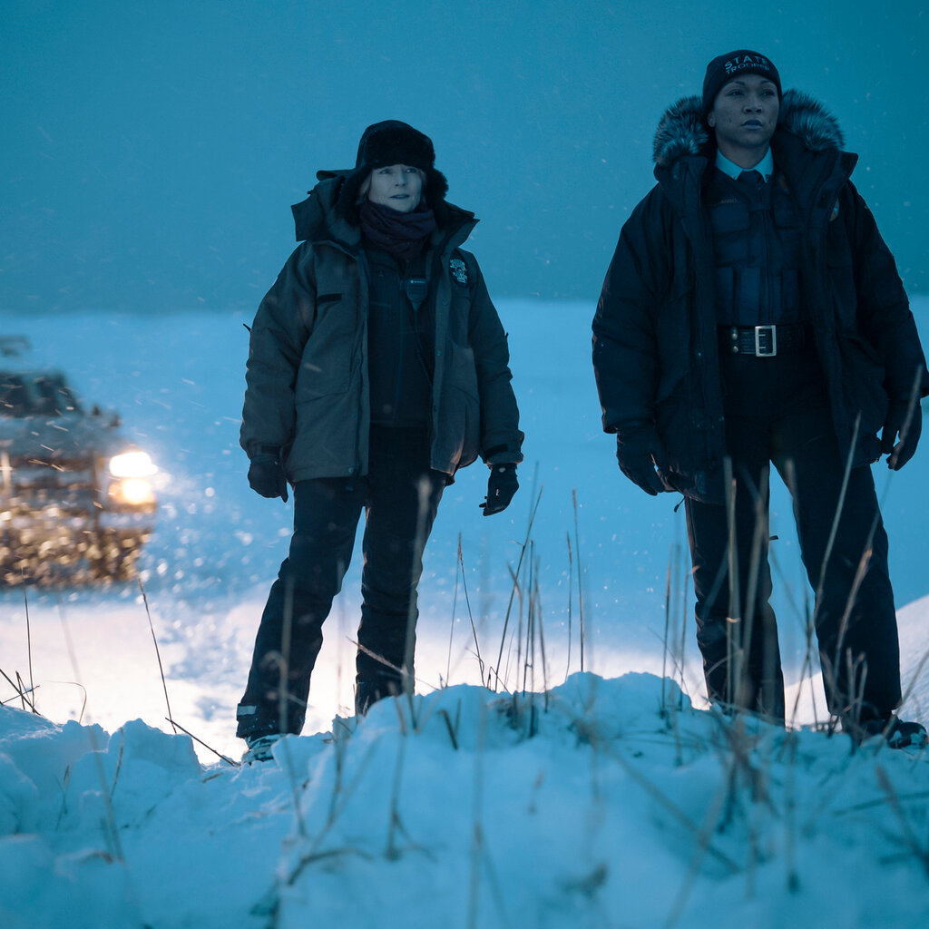 Two female law enforcement officers stand in the snow in dusky light. A police truck sits behind them with the headlights on.