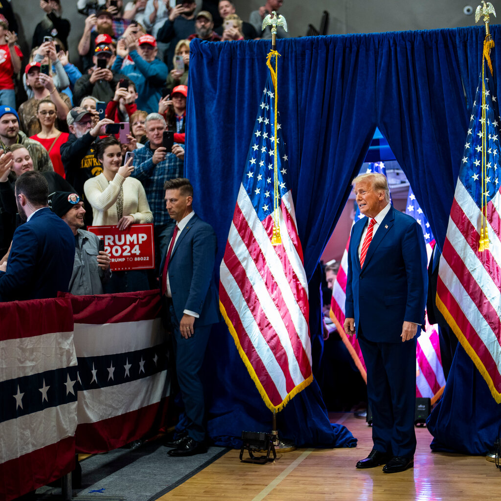 Donald Trump enters a campaign event through blue curtains, with American flags on either side of him. 
