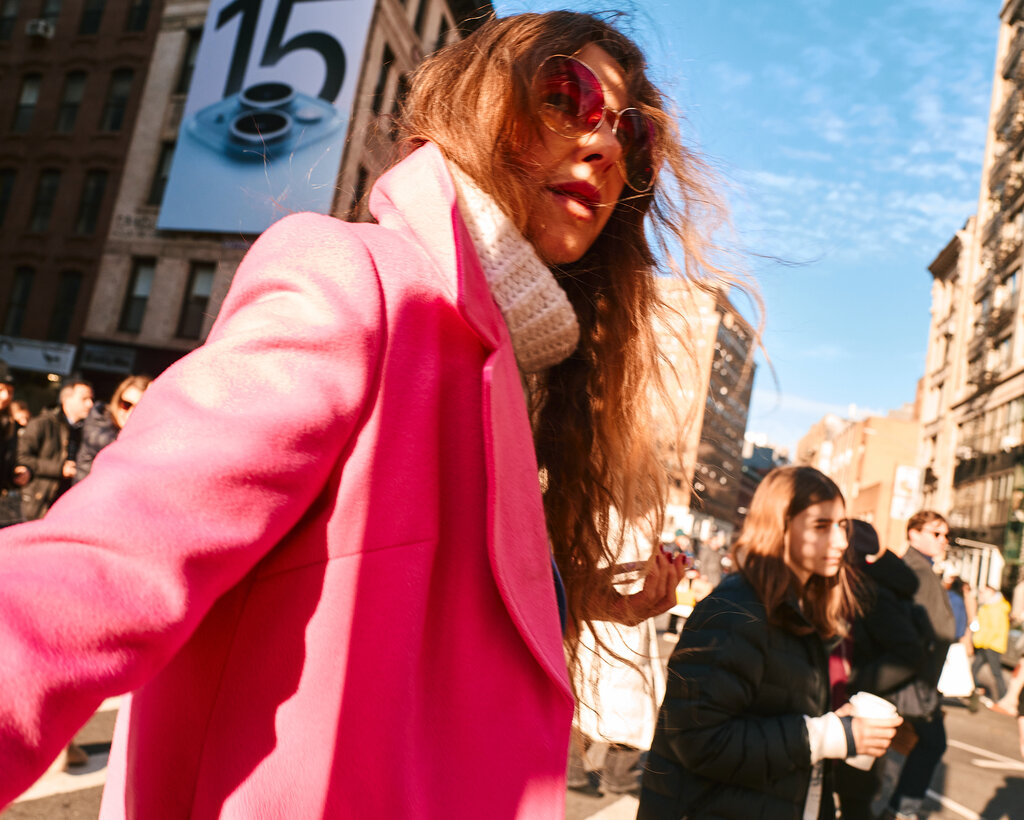 A person, seen from the waist up, walking outside. The person has long brown hair and is wearing sunglasses with pink lenses and a pink overcoat.