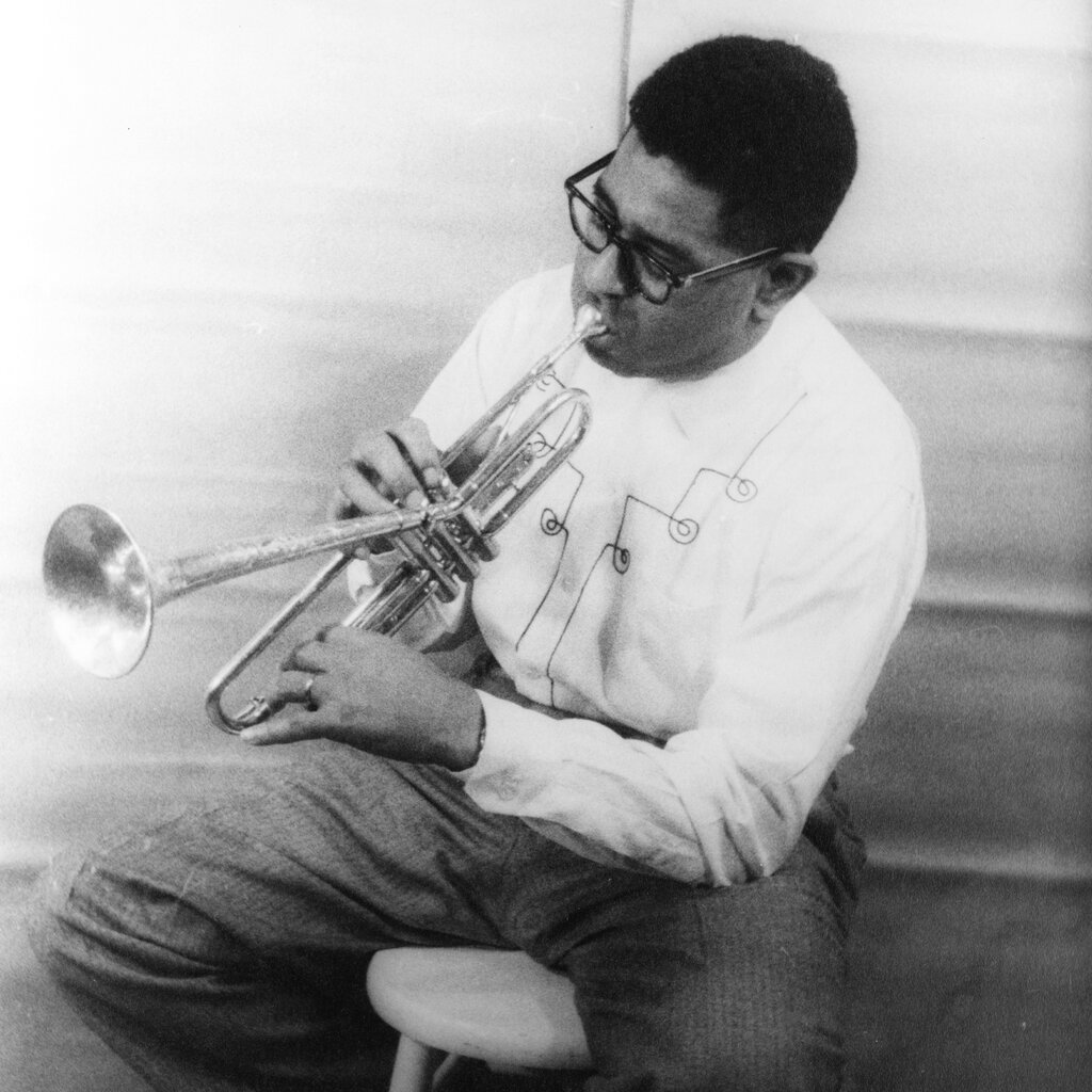 A man sits on a stool and plays an iconic bent trumpet.