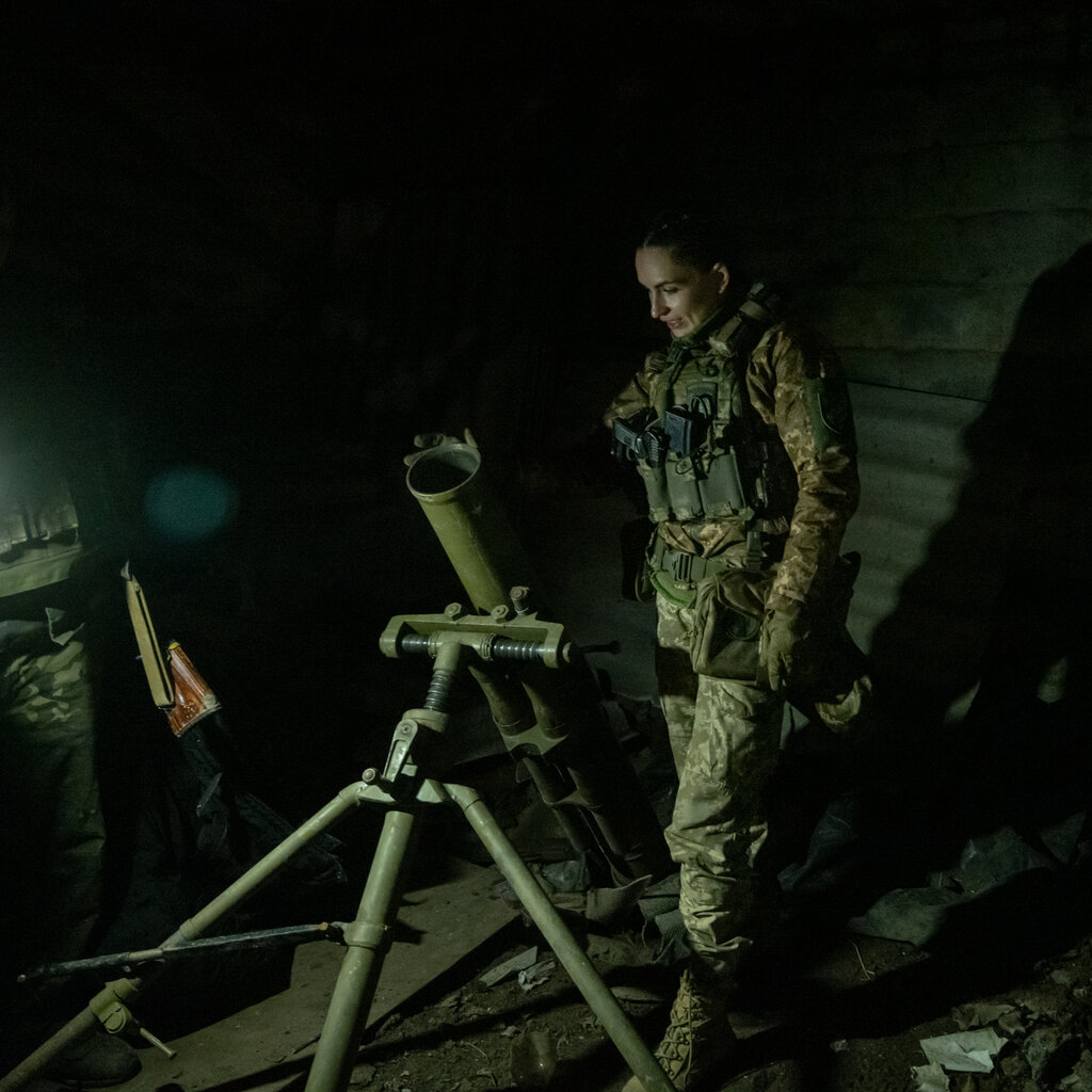 A female soldier wearing camouflage stands behind a piece of military equipment in a dark room.