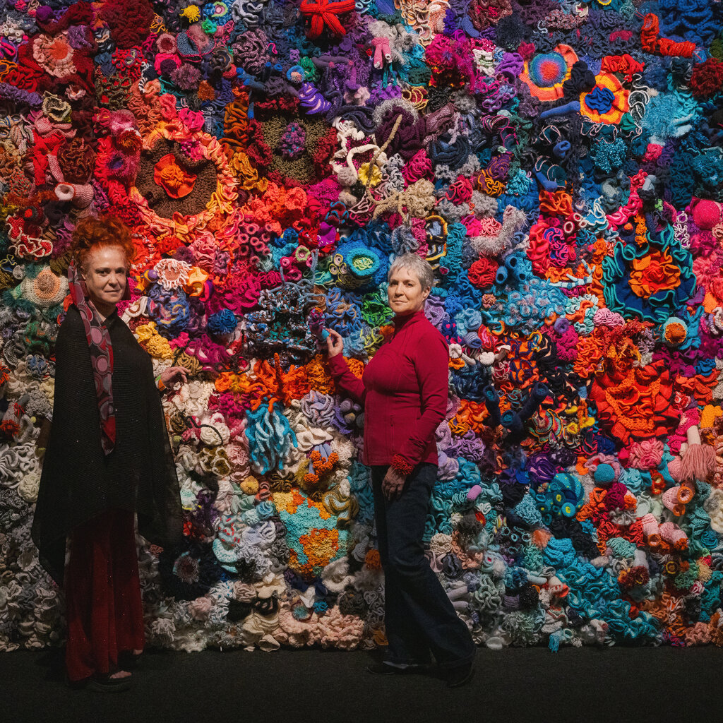 Two women stand in front of a crochet depiction of a coral reef. 