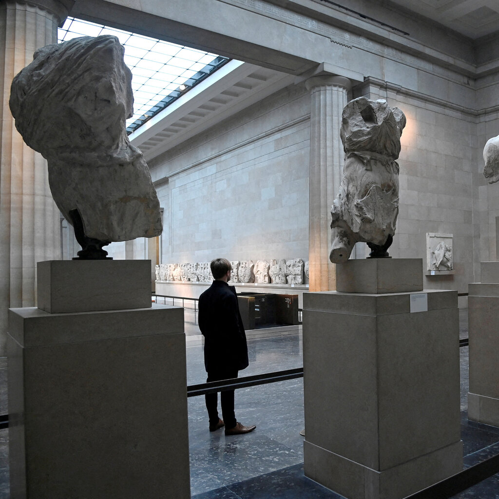 Inside a grand museum space, sculpture fragments are displayed on plinths. 