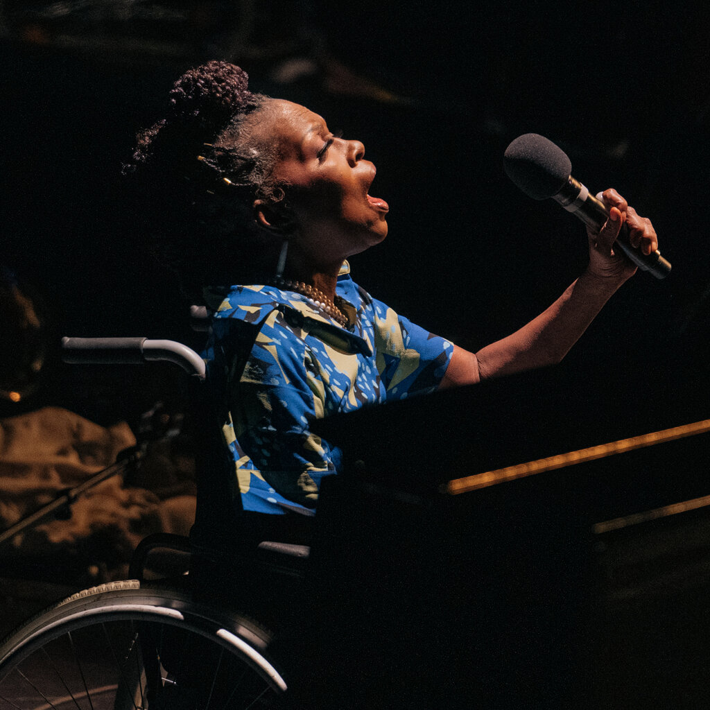A woman in a wheelchair holds a microphone in her left hand, her mouth open as she performs.
