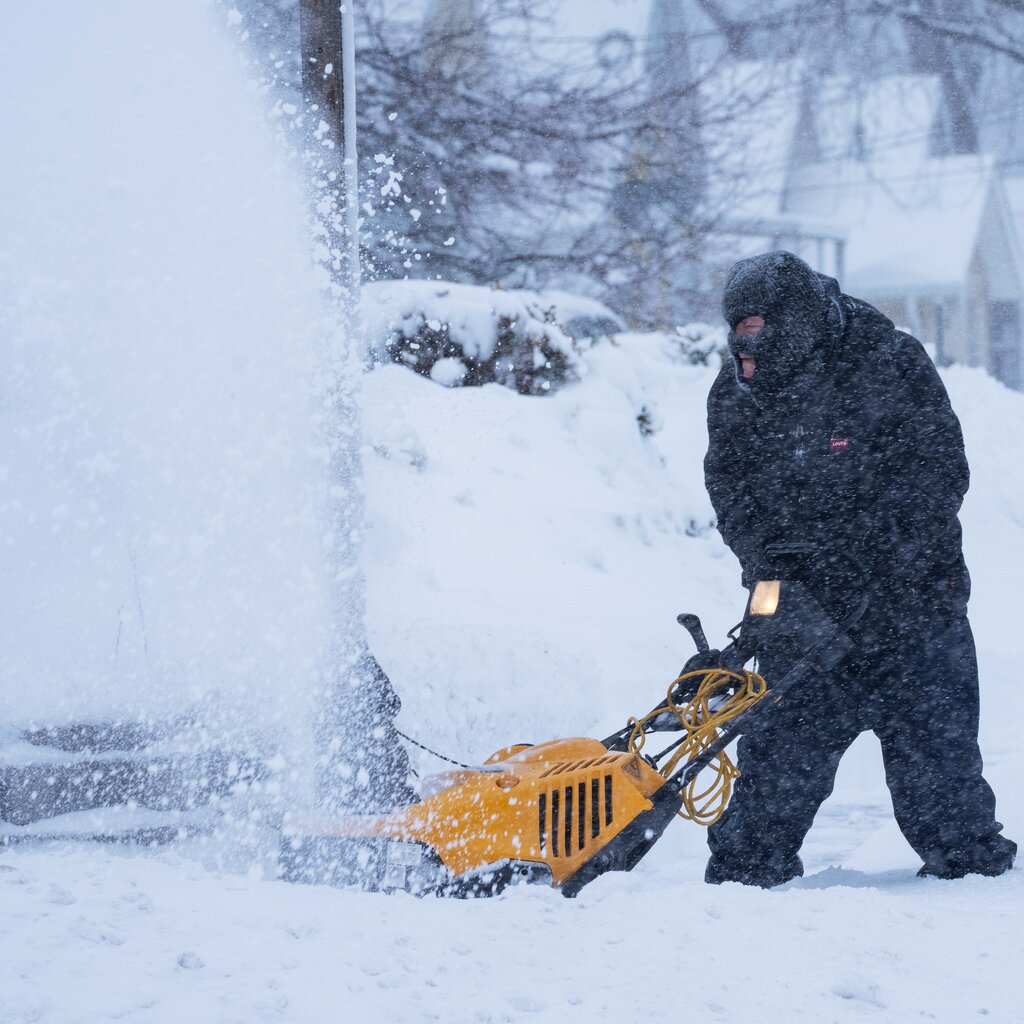 A man pushes a yellow snow plow through knee-high snow. 