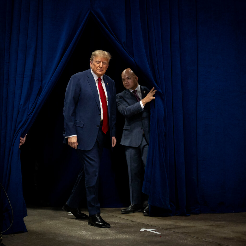 Former President Donald J. Trump in a blue suit. He is walking between blue curtains being held open for him. 