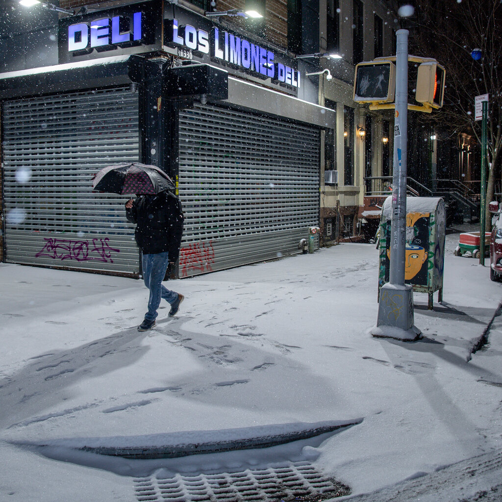 A man carries an umbrella as he walks past a deli on a dark, snow-covered city street.