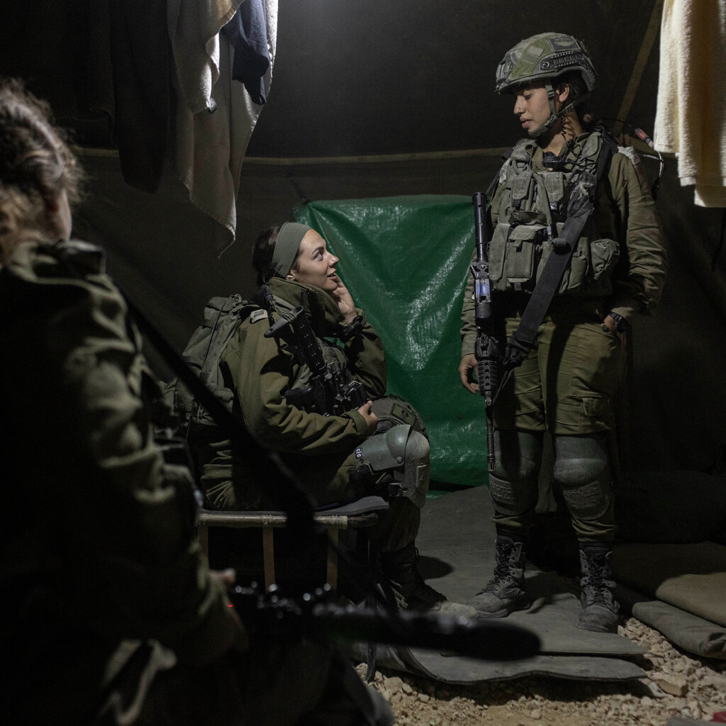Two female soldiers, both in body armor, talking in a dimly lit tent as a third female soldier looks on.