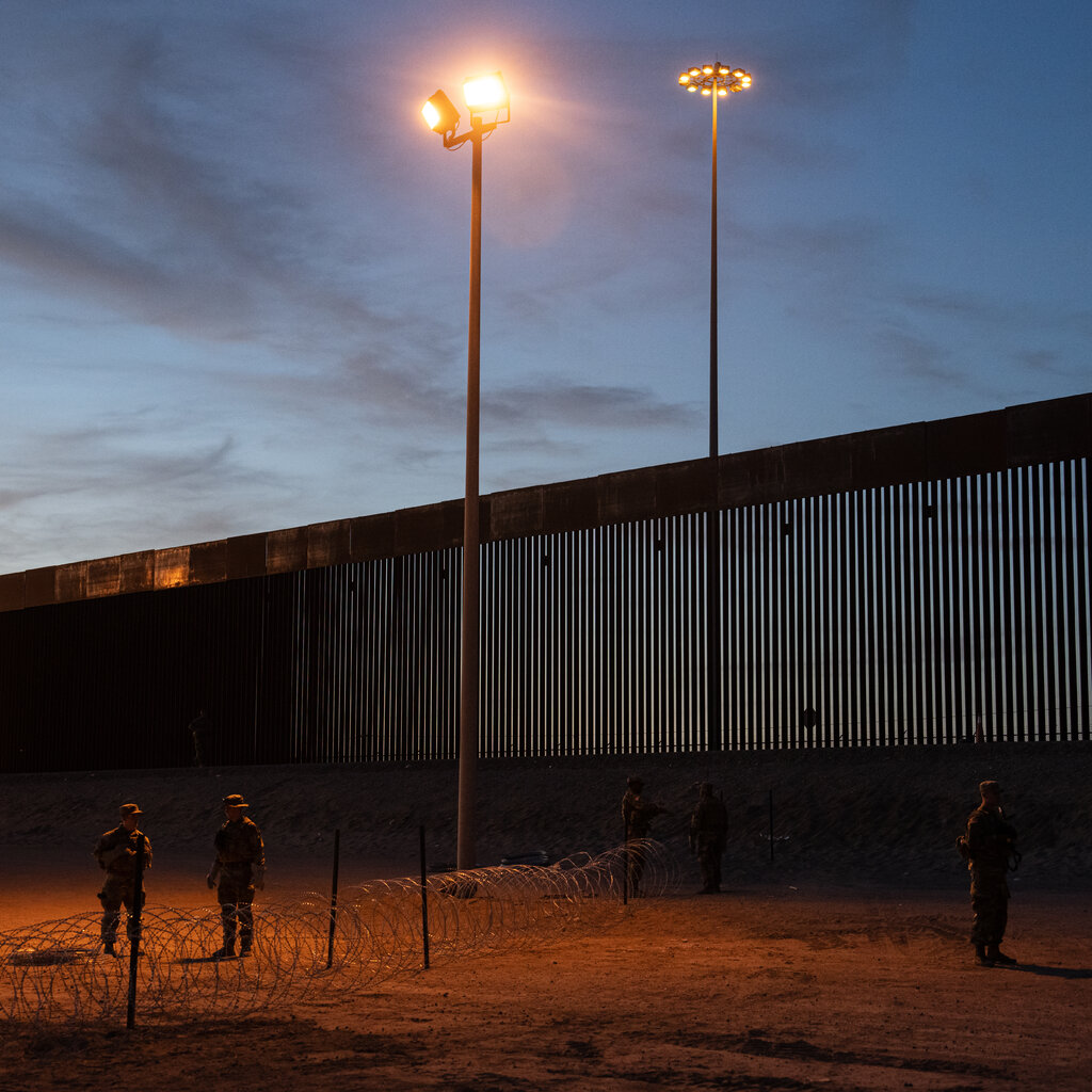 Three National Guard members stand next to a border fence at sunset. In the middle of the image are two light poles. 