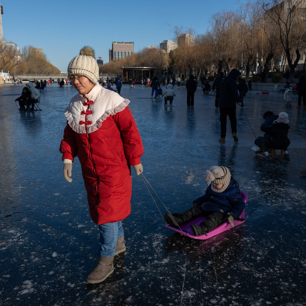 On a frozen river crowded with people, an adult wearing a heavy red coat pulls a child in a pink sled across the ice.
