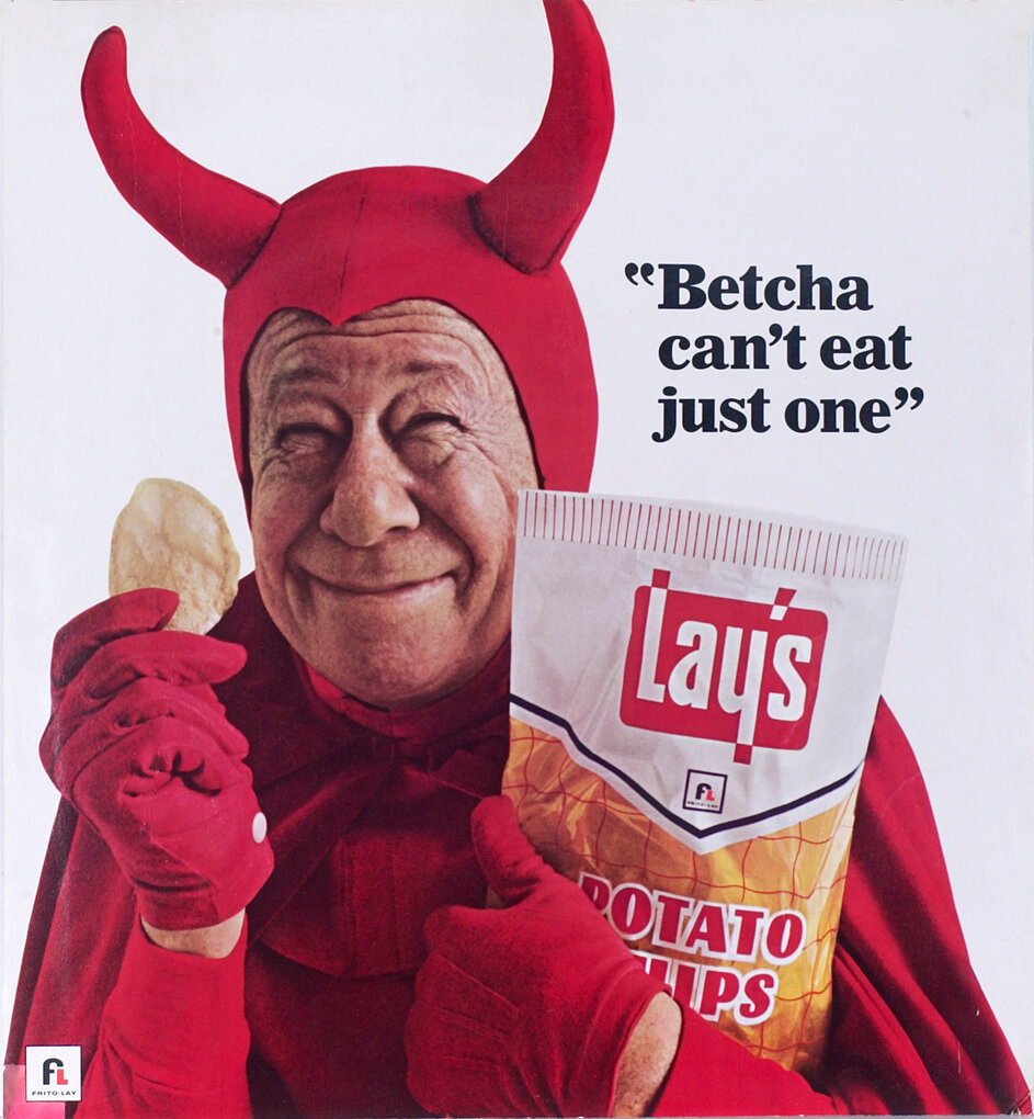 Bert Lahr, smiling in a red devil costume, holds a bag of potato chips next to the words “Betcha can’t eat just one.”