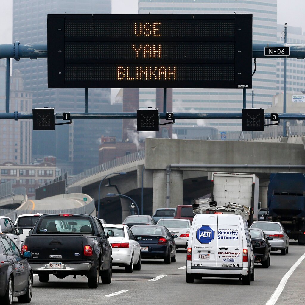 Cars on a busy urban interstate highway drive beneath a safety sign with the message “Use Yah Blinkah.”