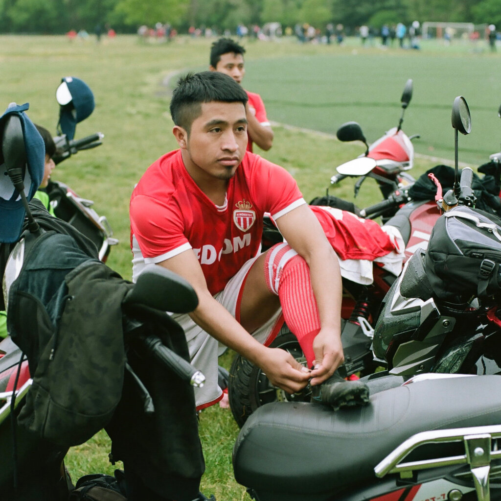 José Estuardo wears a red jersey, white shorts and red socks. He ties his shoe as it rests on a scooter. In the background people stand on a soccer field.