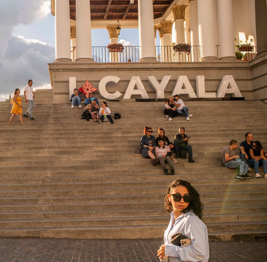 People sitting on steps leading up to a balcony with fancy columns. Large letters at the top of the steps spell “I” followed by a flowerlike symbol and the word “Cayala.”  