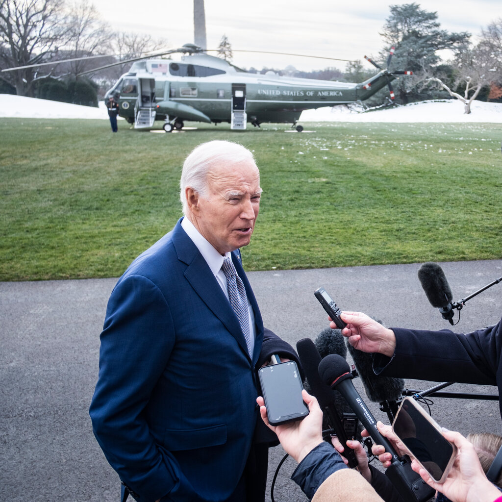 President Biden, in a blue suit, speaking with reporters. A helicopter is behind him.