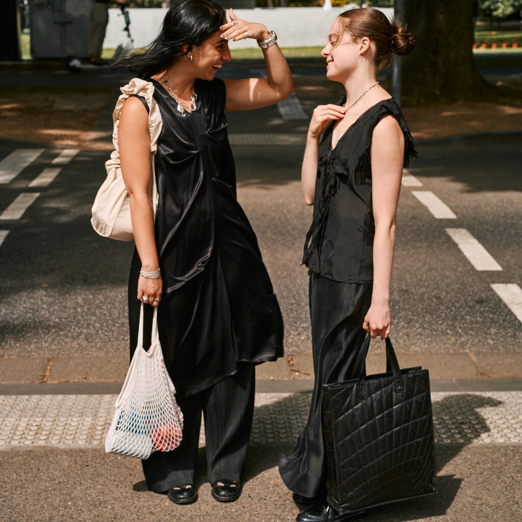 Two people talking to each other on a city street. One person, left, has long dark hair and is wearing a shiny black sleeveless dress over dark pants and black shoes. The other person has brown hair styled in a bun and is wearing a black sleeveless top, black pants and black shoes.