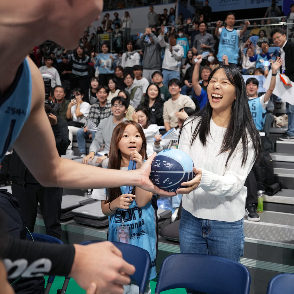 A male player in a sports uniform hands a blue basketball to a woman. Other fans who surround her are cheering.
