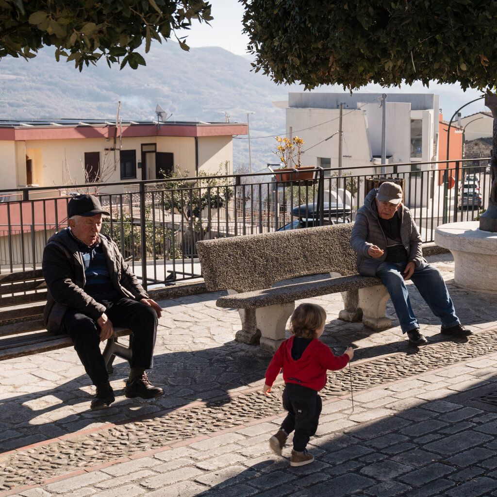 Two older men sit on benches in a town square. A toddler runs past them. 