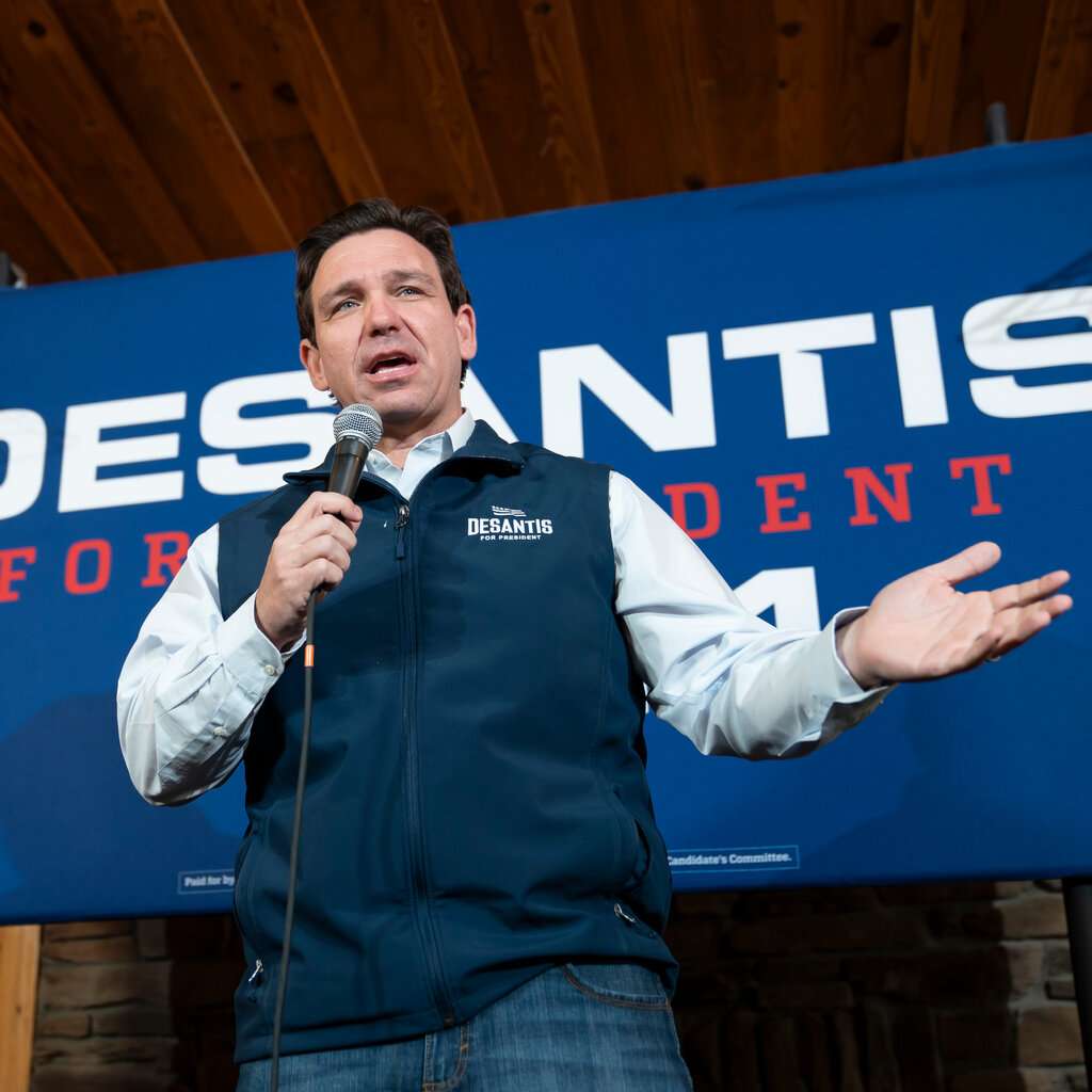 Ron DeSantis, dressed in a navy campaign zip-up vest with his name on it, stands on stage speaking into a microphone. A poster with the words "DeSantis for President" in the background. 