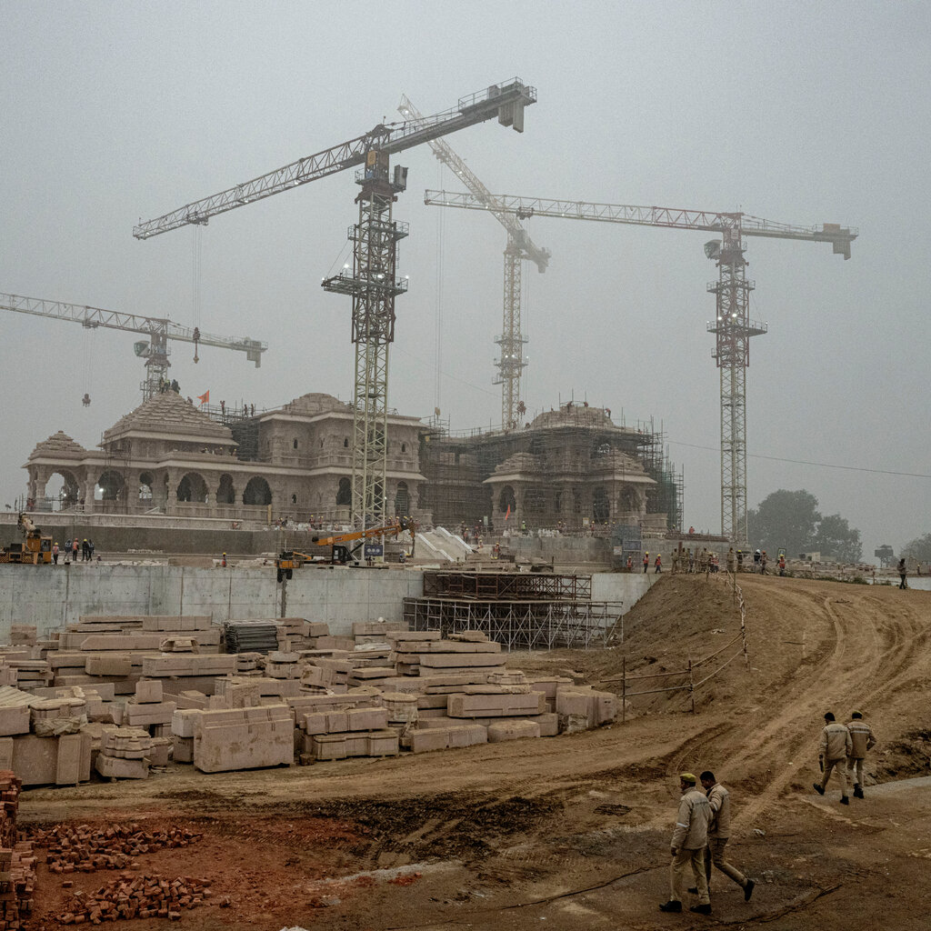 Workers walk near a large temple complex that is under construction. Cranes loom over the project, and large slabs of stone are piled nearby. 