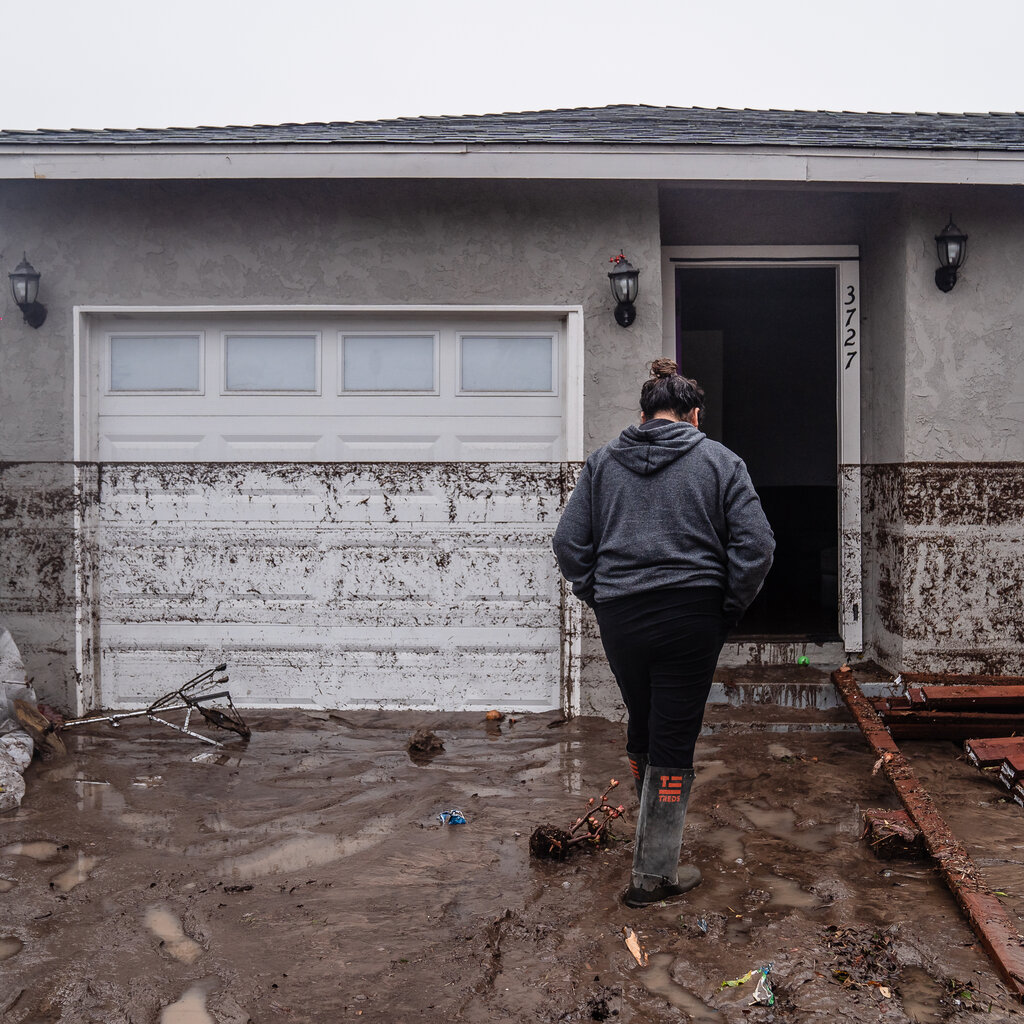 Celene Pulido walks up to her house wearing rubber boots as mud covers her driveway. A layer of mud residue is about three feet high on her exterior walls and garage.