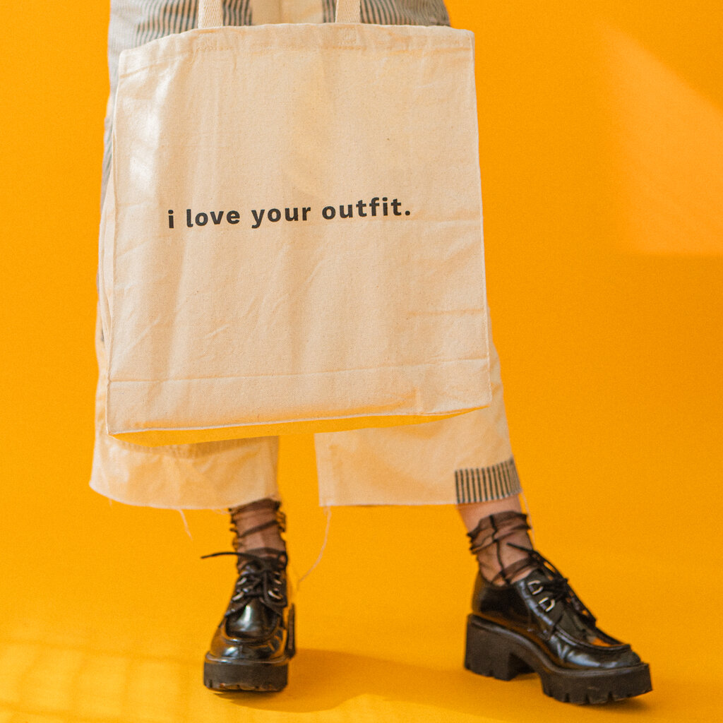 A close up of a woman’s chunky black loafer-esque shoes with heels, and sheer black socks. She wears wide leg pants and holds up a white tote bag with the words, “I love your outfit” in black lettering. 