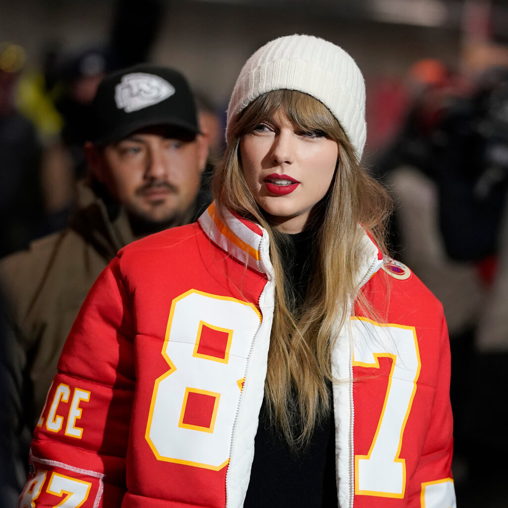 Taylor Swift, wearing a white knit hat and a red jacket with No. 87 on it, walks through a crowd.