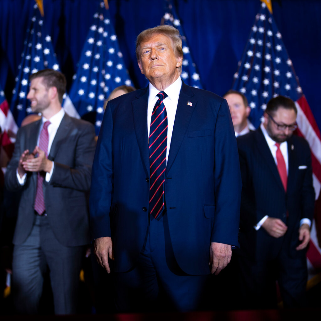 Donald Trump stands on stage in a suit and tie. Behind him, out of focus, are American flags and a group of people. 